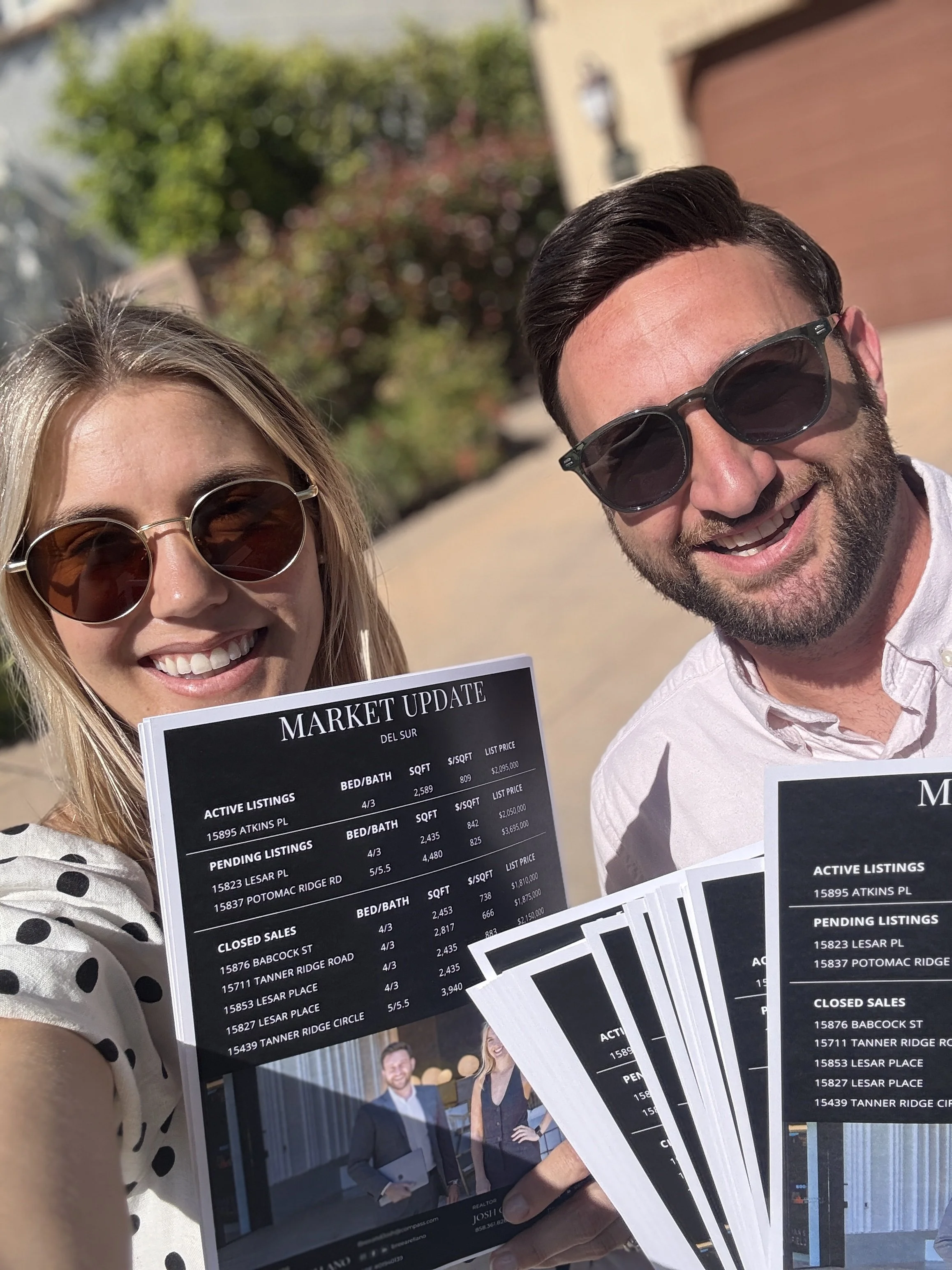 A woman and man smiling while holding real estate market update flyers outdoors on a sunny day.