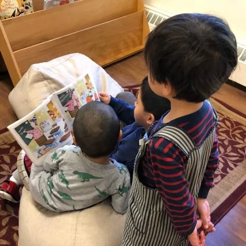 Three children, one sitting on a bench and the other two standing, are looking at a picture book together. The setting appears to be indoors in a cozy room.