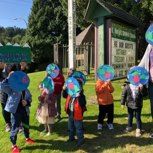 Group of children holding paintings of the earth outside near a sign and a green fence.
