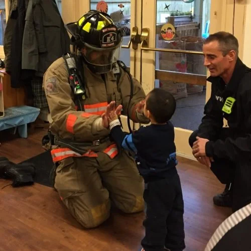 Firefighter in full gear kneeling and giving high five to young boy inside a building, with another firefighter squatting nearby.