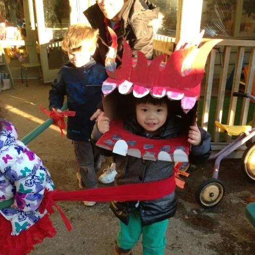 Two young children outdoors, one wearing a Chinese dragon costume with a large, colourful head and open mouth, smiling and holding its head.