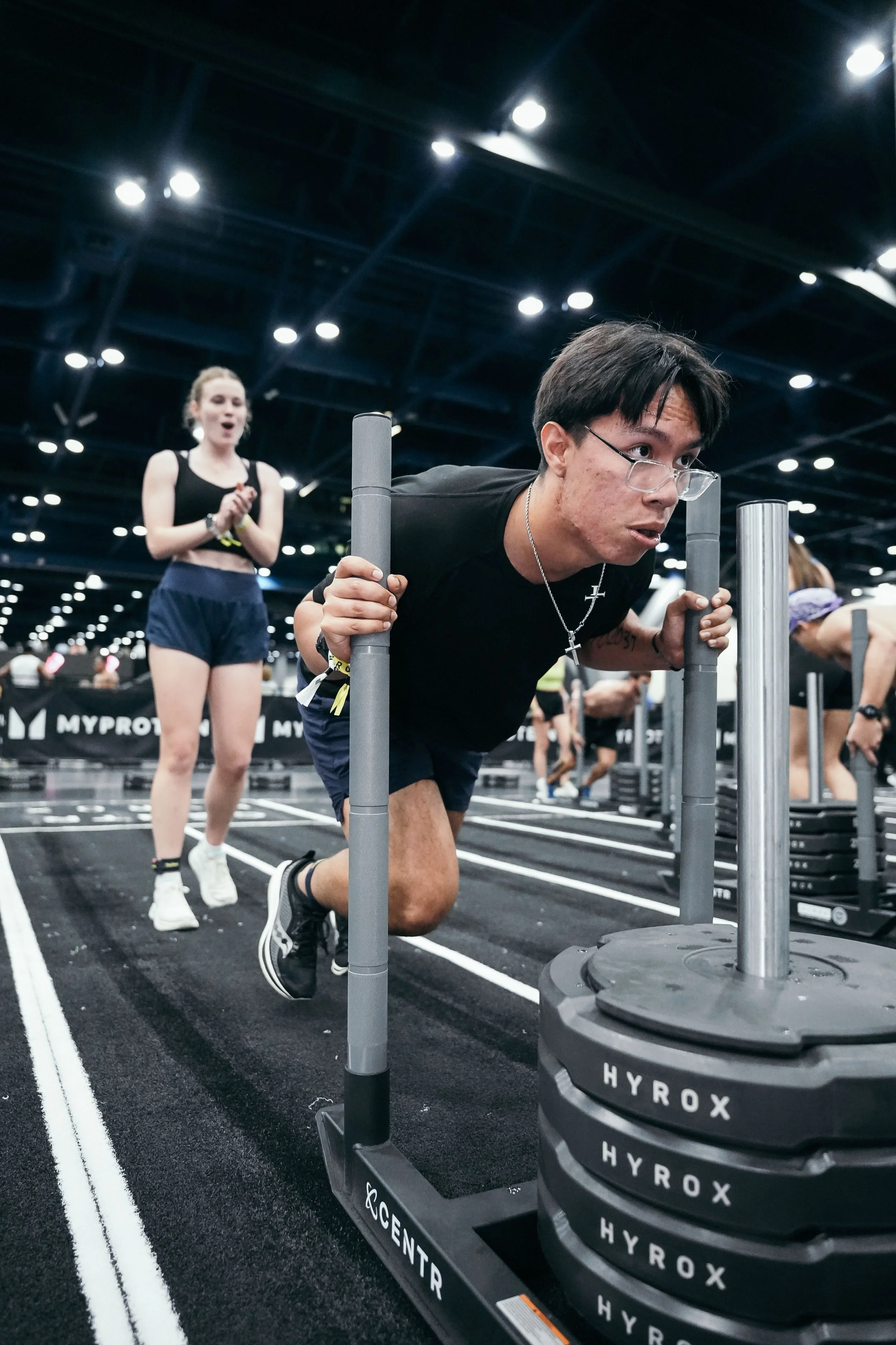 A man pushing a weighted sled at a gym with a woman clapping in the background.