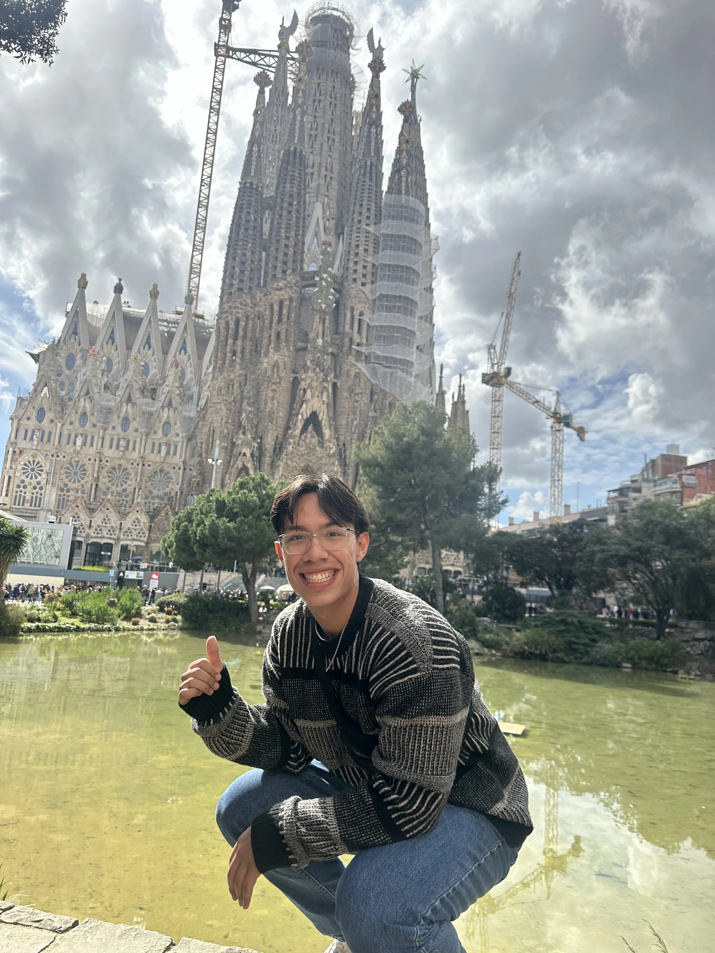 A smiling young man crouching near a pond with the Sagrada Família basilica in Barcelona in the background, under partly cloudy skies.