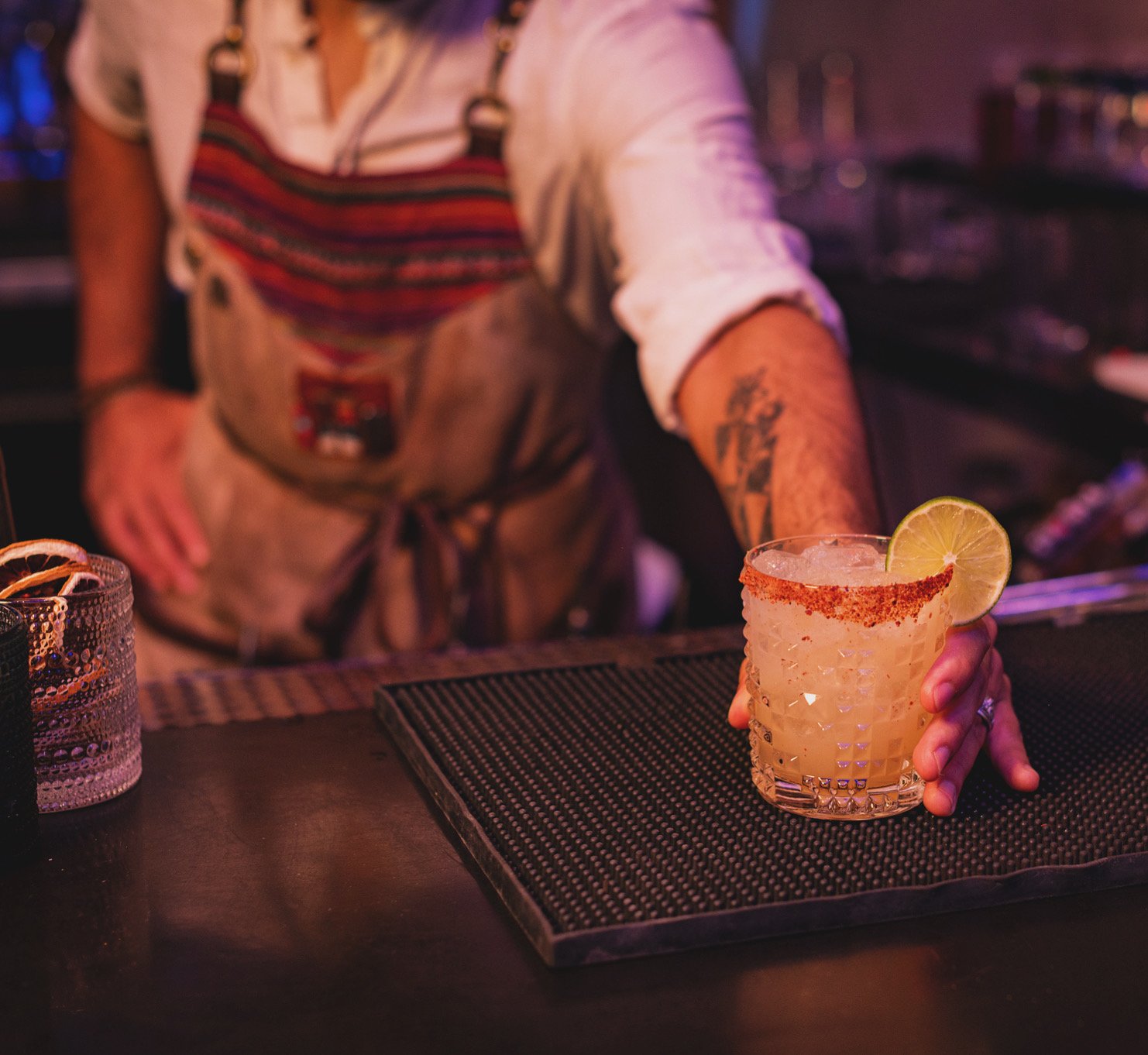 A bartender presenting a cocktail garnished with a lime wedge at a bar, with a tattooed arm and apron.