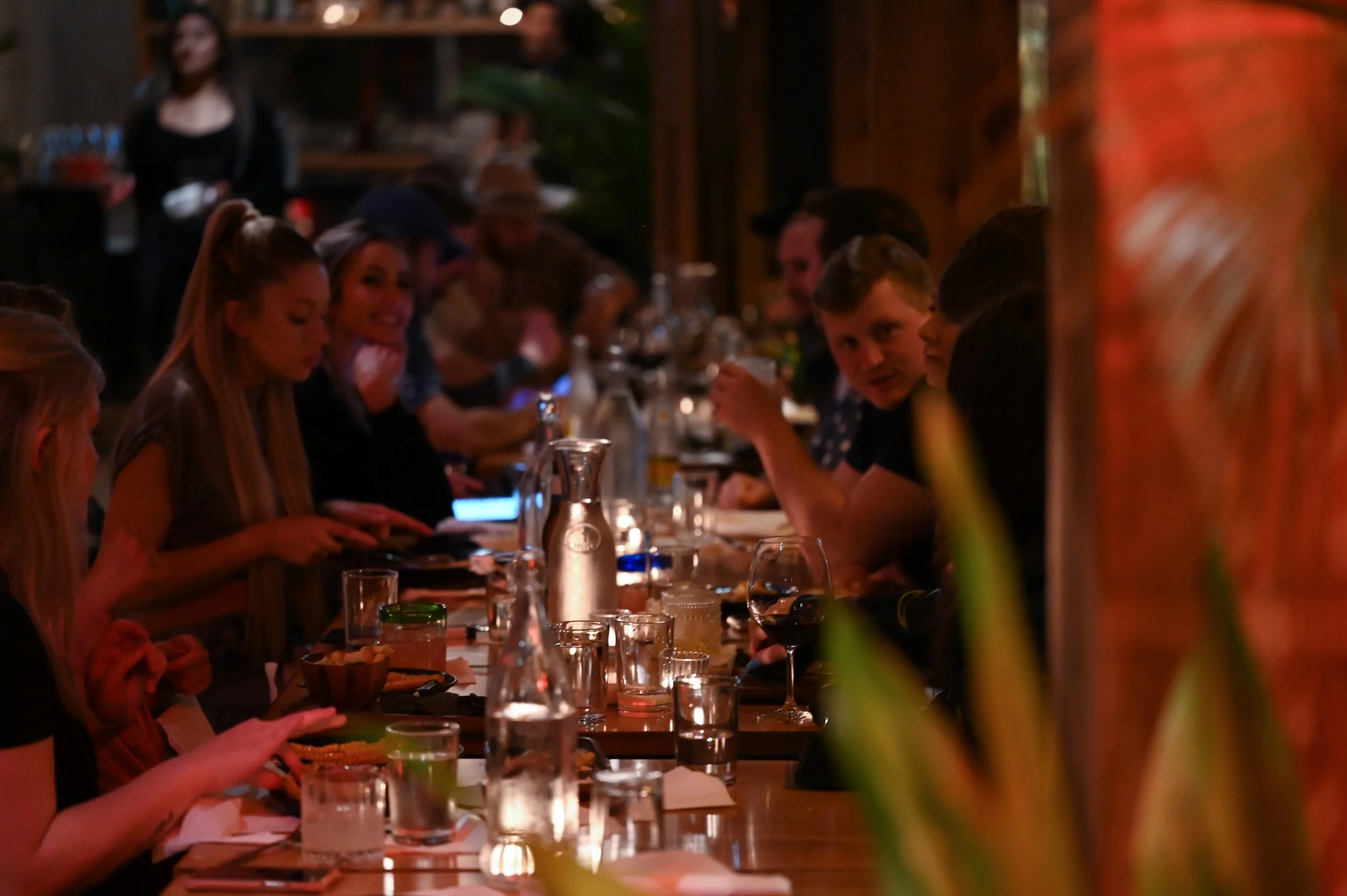 People sitting at a long table in a dimly lit restaurant, with glasses, bottles, and plates, some engaging in conversation and others looking at their phones.
