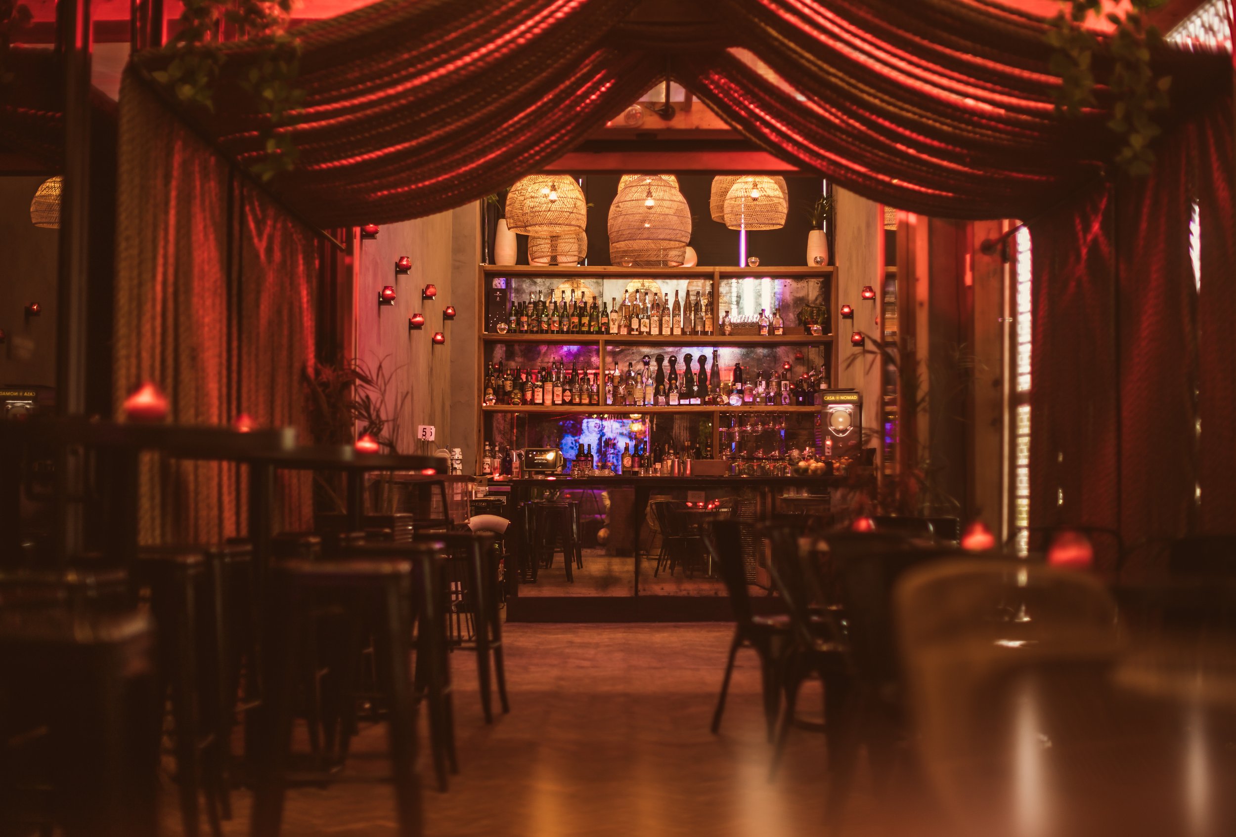 A dimly lit bar with warm red and orange tones, decorated with hanging woven lamps and red drapes. Shelves behind the bar are stocked with bottles of liquor, and small red candles are placed along the sides of the seating area.