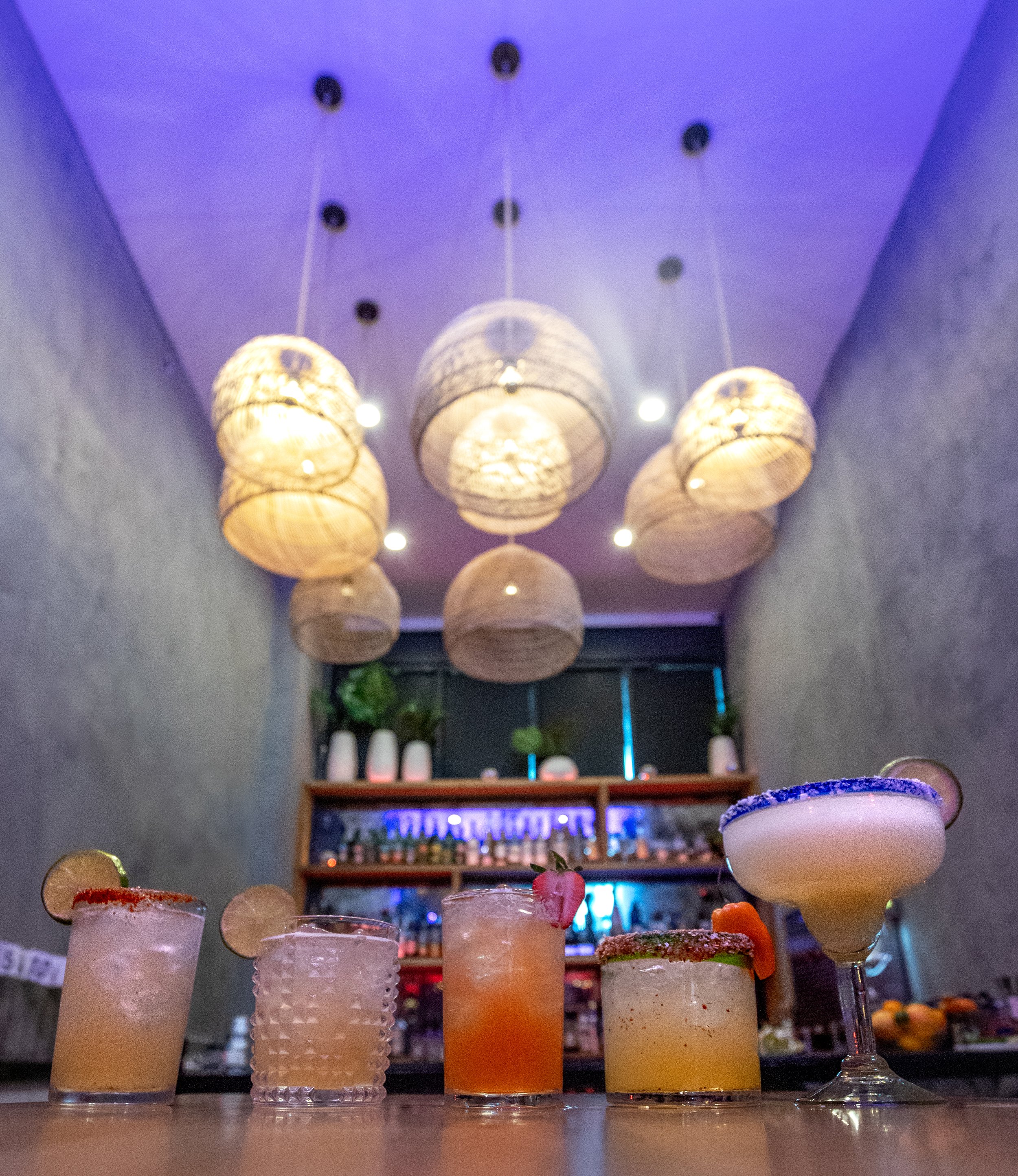 Five colorful cocktails on a bar counter in front of a shelf with bottles, with hanging woven lantern lights and a purple-lit ceiling.