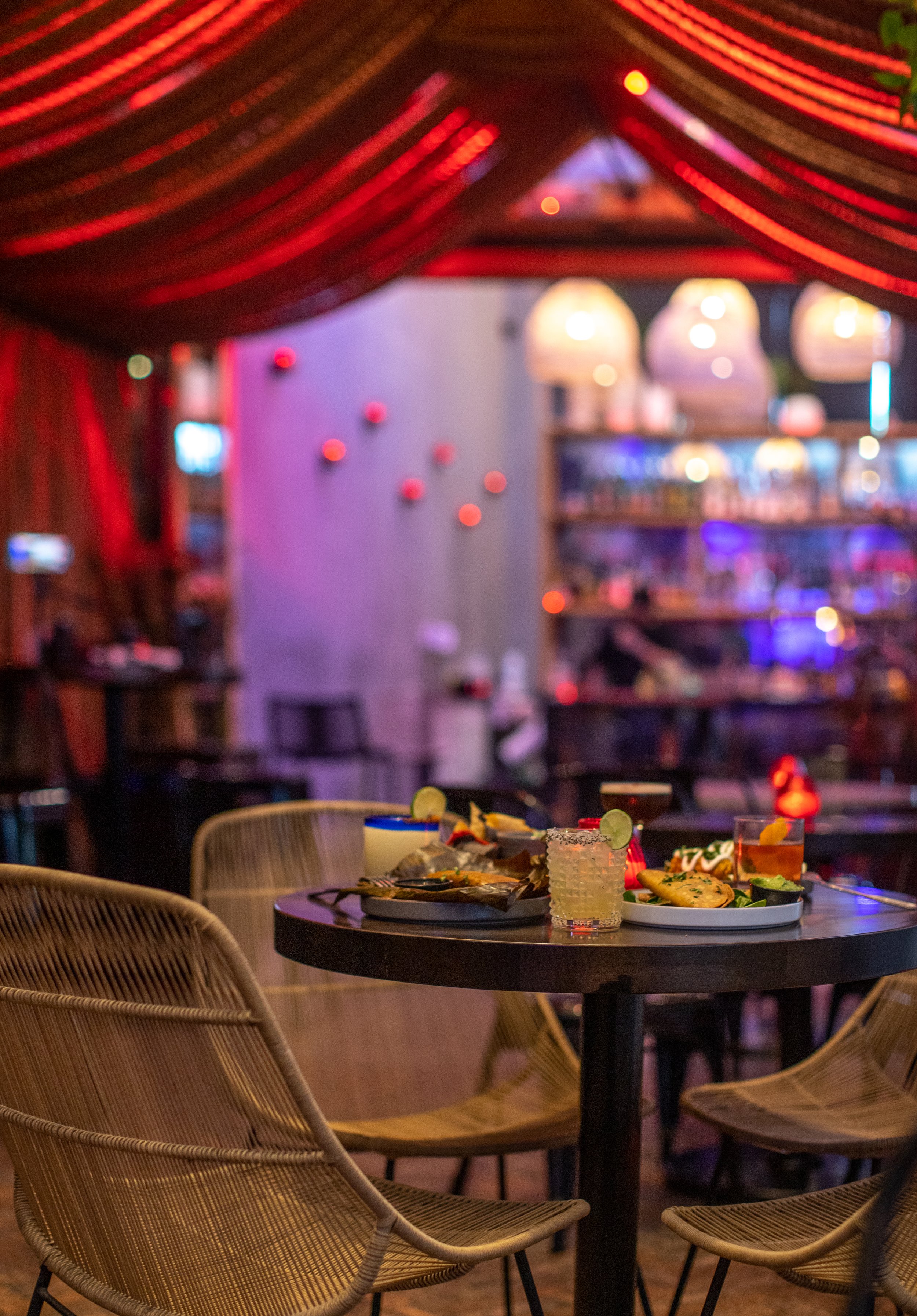 A round table in a restaurant with various plates of food and drinks, surrounded by wicker chairs, with colorful lighting and blurred background.