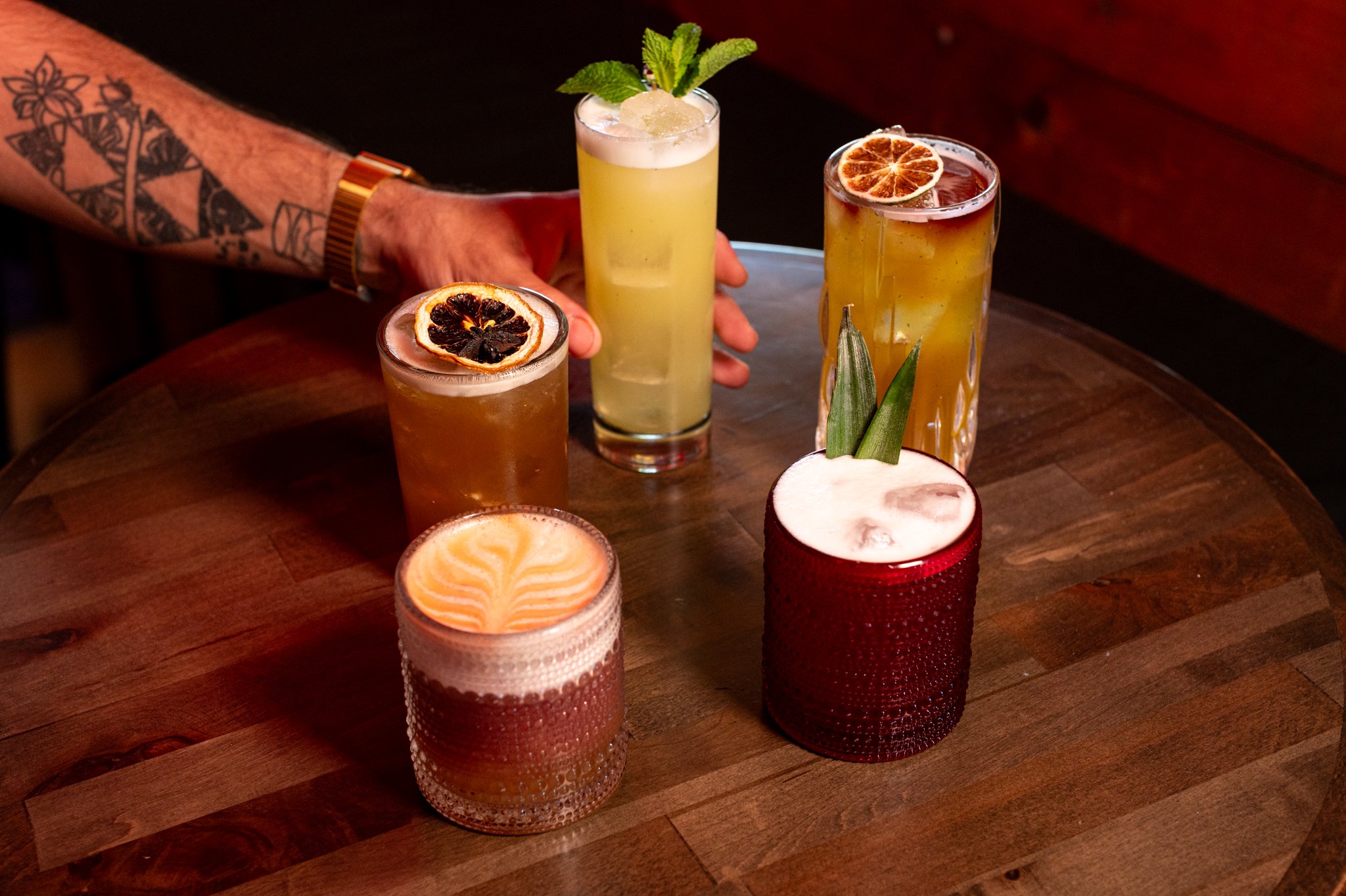 A man with tattoos and a gold watch holding a tall glass of yellow cocktail garnished with mint leaves, surrounded by five other drinks with fruit garnishes on a wooden table.