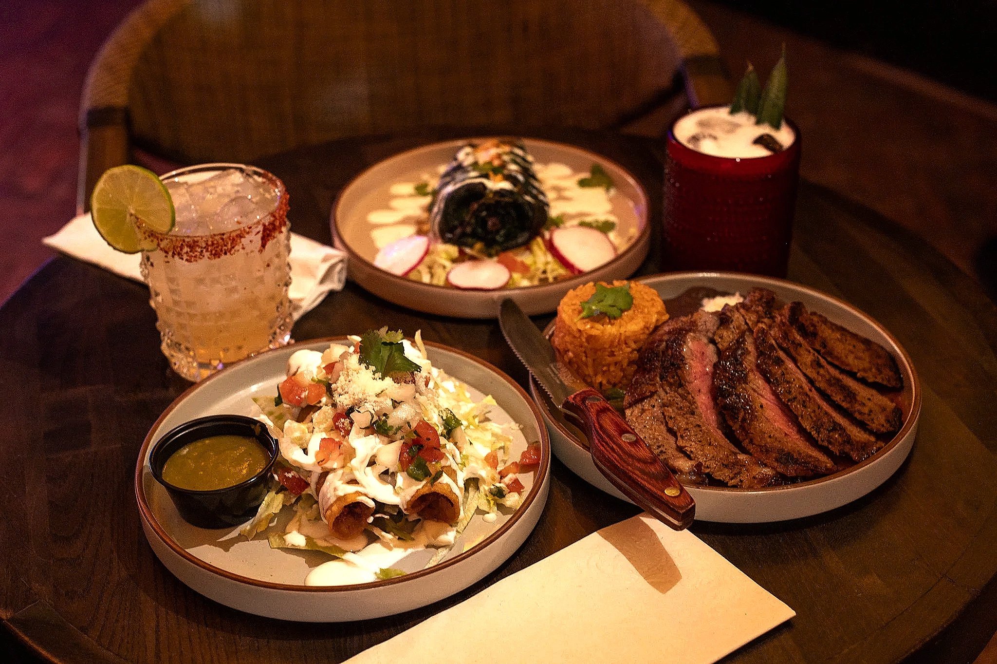 A Mexican meal with grilled steak, Mexican rice, a salad, and a rolled taco on a wooden table, accompanied by a lime margarita and two other drinks.