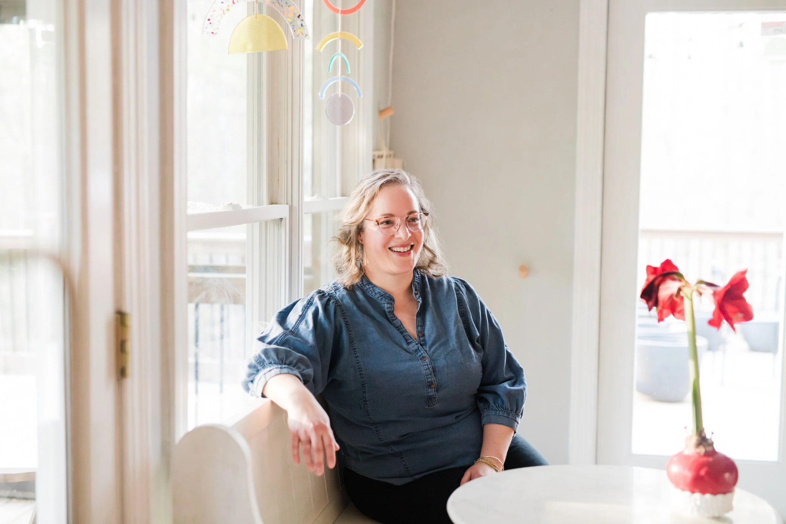 A woman with curly blonde hair and glasses, wearing a denim shirt, smiling and sitting near a window in a bright room with pastel-colored hanging decorations.