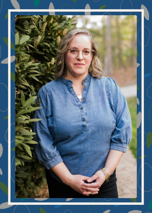 A woman with light brown hair and glasses standing outdoors next to green foliage, wearing a denim shirt and black pants.