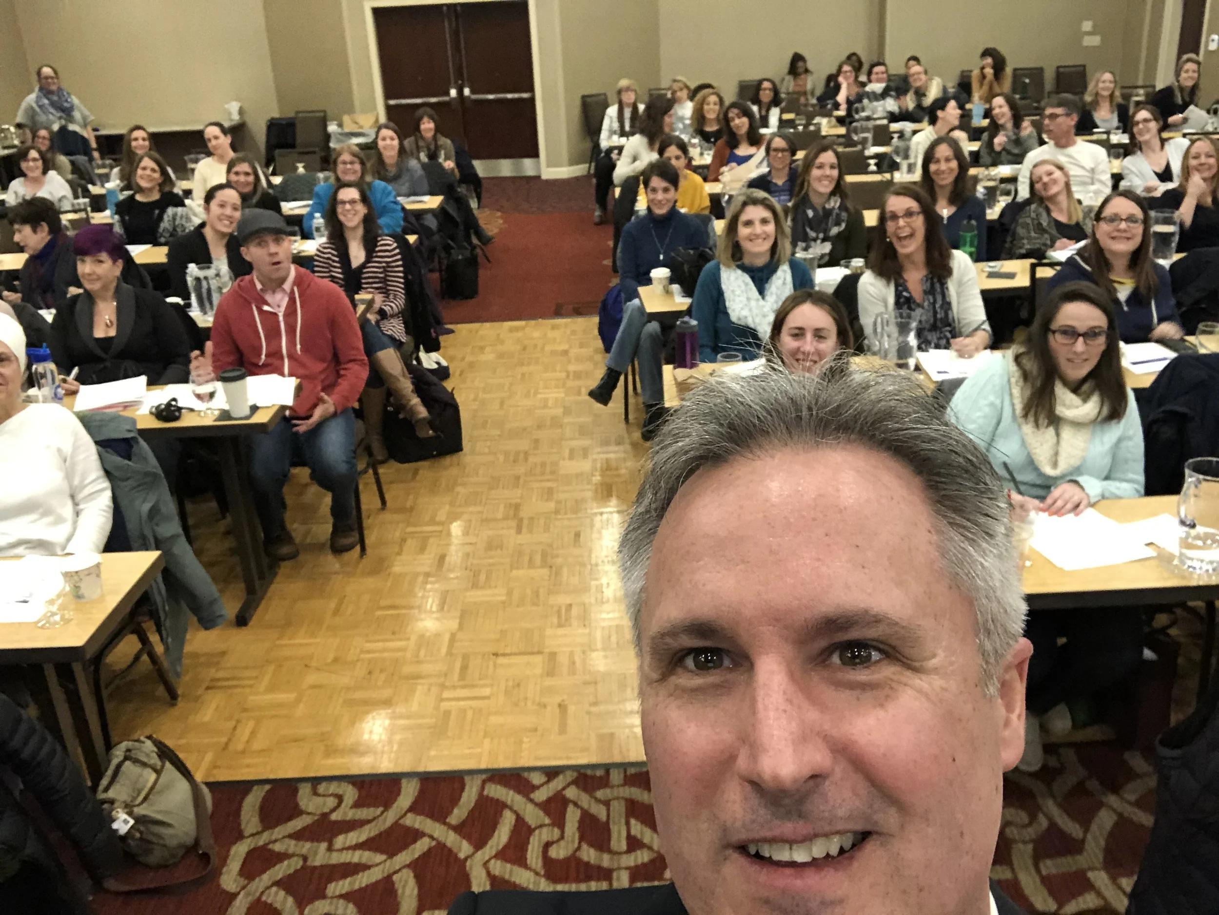 A group of people attending a conference or seminar, sitting at tables in a large room, with a man taking a selfie in the foreground.