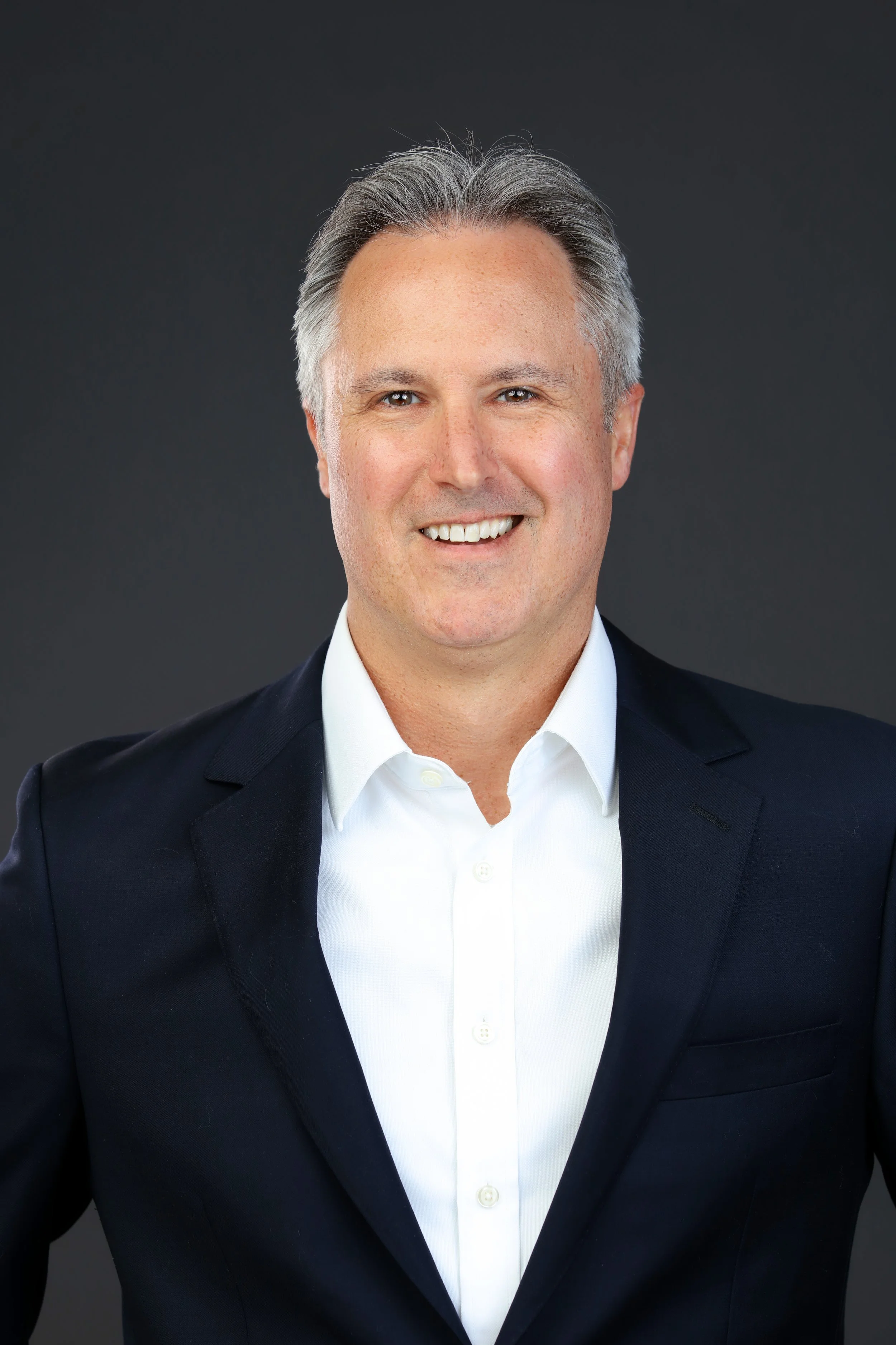 Headshot of a smiling man with gray hair in a white dress shirt and dark blazer against a dark gray background.
