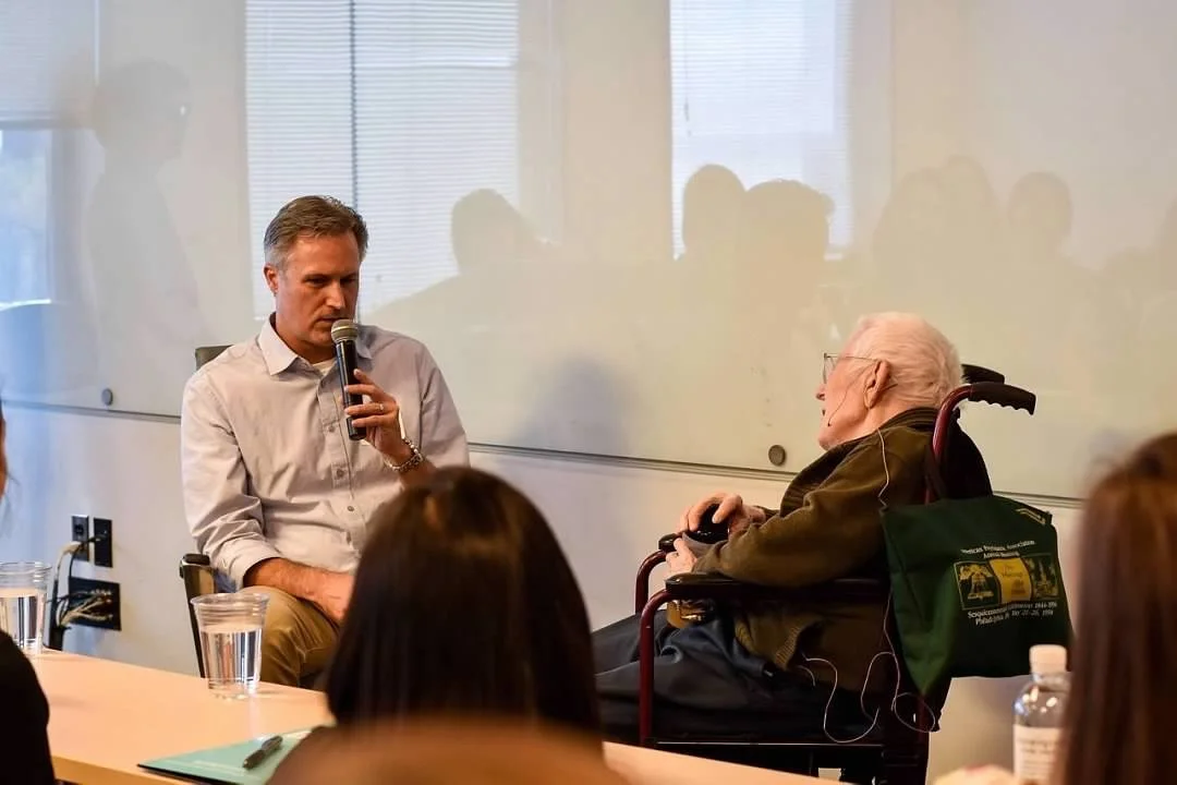 A man holding a microphone sits at a table in a conference room, speaking to an elderly man in a wheelchair. Several people are seated around the table, listening. There are glasses of water, notebooks, and a water bottle on the table, with a large window and a whiteboard in the background.