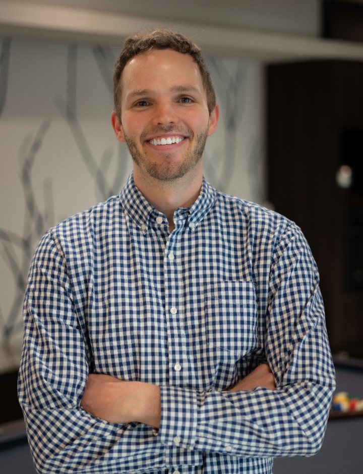 A smiling man with crossed arms standing indoors, wearing a blue and white checkered shirt.