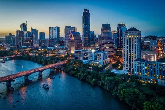 Skyline of downtown city with tall buildings, river with boats, and a bridge at sunset.