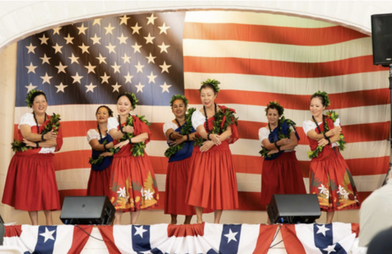 Group of women in traditional Hawaiian attire performing a dance on stage with American flag backdrop.