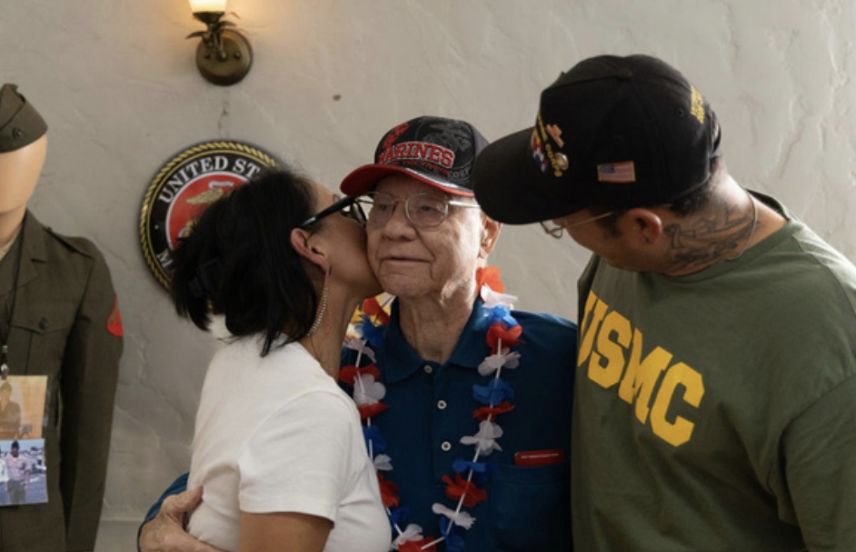 A woman is kissing an elderly man on the cheek, both smiling. A man wearing an 