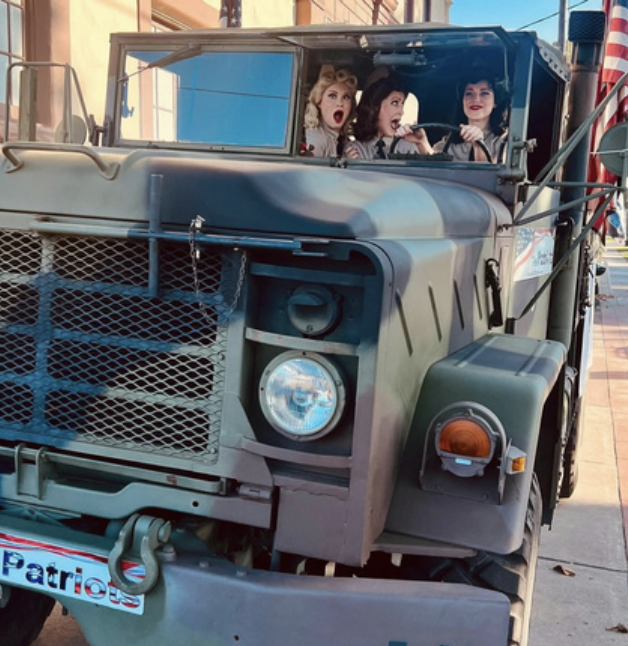 Three women riding in the front of a military-style vehicle, making expressive faces, as they pass by a storefront with American flags and patriotic decorations.