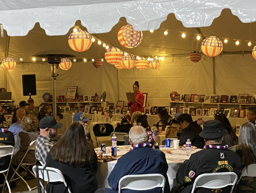People sitting at round tables inside a large white tent, decorated with red, white, and blue paper lanterns and string lights, attending a patriotic event where a woman is speaking on stage, with framed photos and artwork displayed behind her.