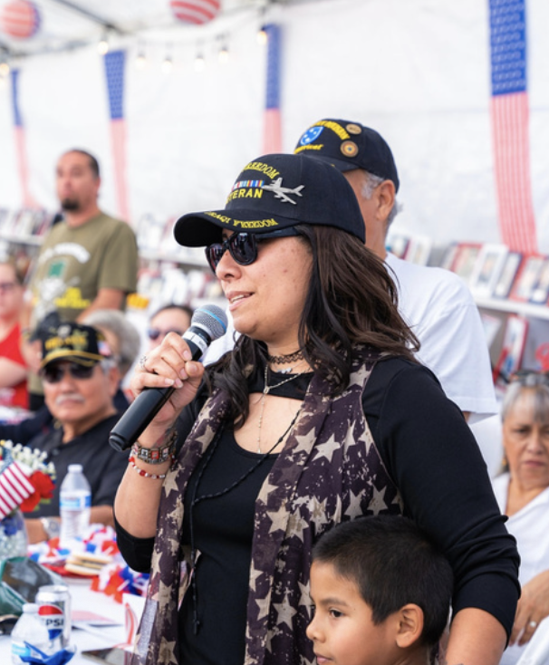 A woman wearing sunglasses and a navy cap with patriotic patches, speaking into a microphone at a patriotic event, surrounded by older adults and children, with American flags and patriotic decorations in the background.