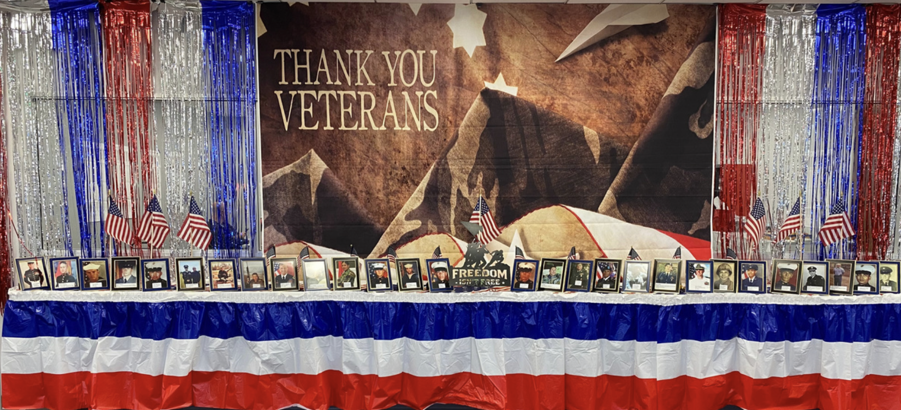 Memorial display for veterans with patriotic decorations, American flags, framed photographs of service members, and a large backdrop with 'Thank You Veterans' written on it.