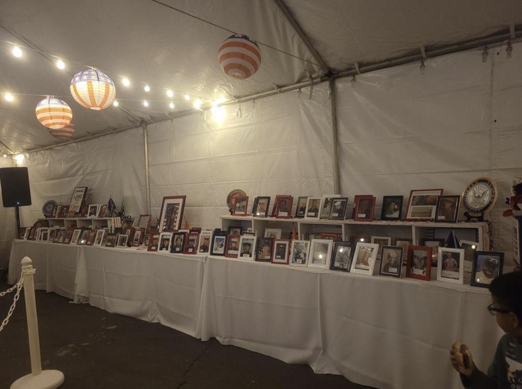 Display of framed photographs and memorabilia at a patriotic event under a large tent with American flag-themed lanterns and string lights.