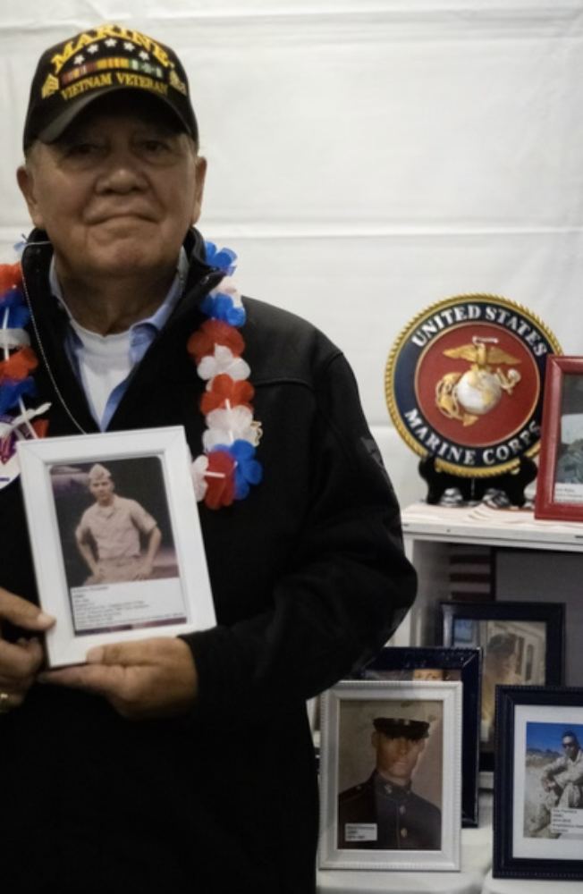 Man wearing a black cap and jacket, holding a framed photo, standing in front of a United States Marine Corps emblem and display of framed photos.