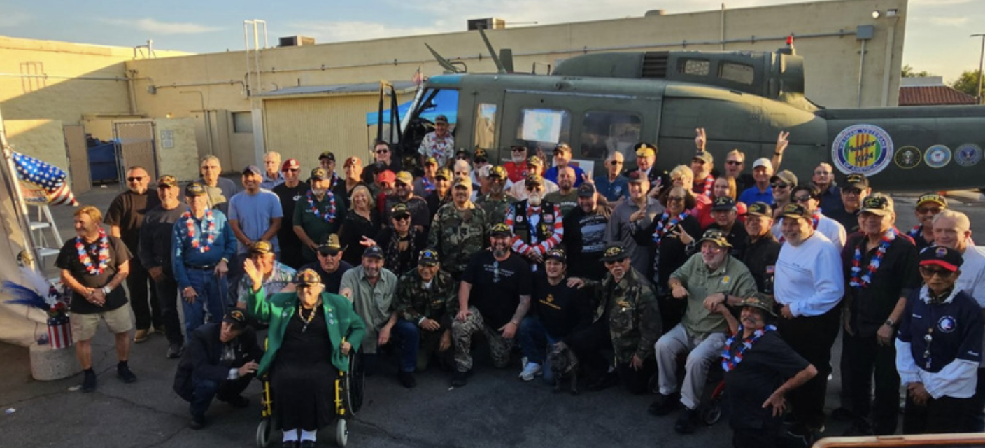 Group photo of people gathered outdoors near a military helicopter, some individuals are wearing patriotic accessories and military uniforms.