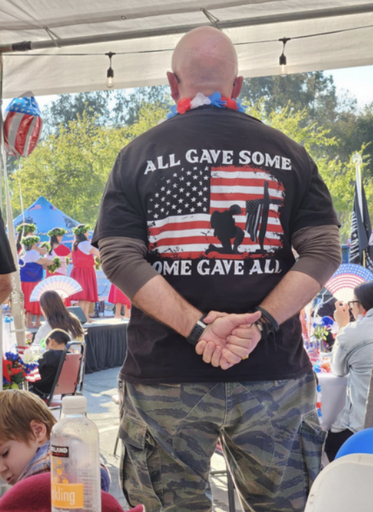 A man stands with his back to the camera at an outdoor event, wearing a patriotic-themed T-shirt with an American flag and a silhouette of a soldier, with words 'All Gave Some, Some Gave All.' There are people, decorations, and trees in the backgroun