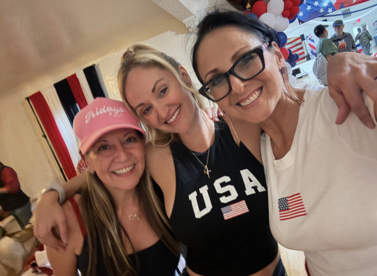 Three women smiling and hugging, celebrating a patriotic event with American-themed decorations in the background. One wears a pink hat with 'Dixie' written on it, another wears a USA tank top, and the third has a USA flag sticker on her white shirt.