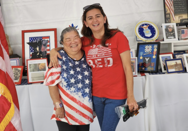 Two women smiling and hugging at an American-themed event, with patriotic decorations and framed photos on display behind them.