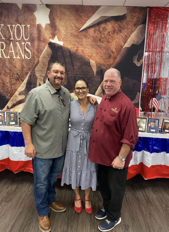 Three people standing together at a patriotic event with a flag-themed table and framed photos in the background.