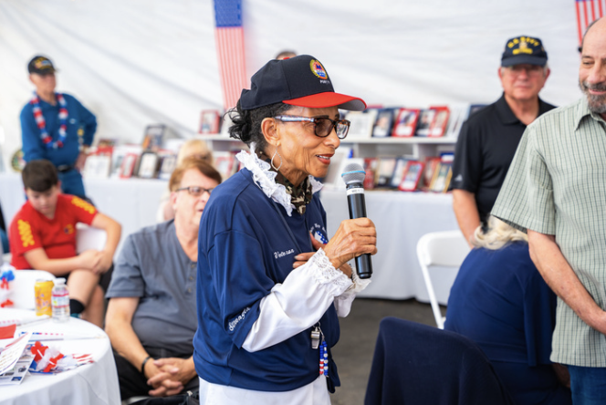 An elderly woman with glasses, wearing a navy blue cap and shirt, holding a microphone and speaking at a gathering in a tent with American flags and framed pictures in the background.