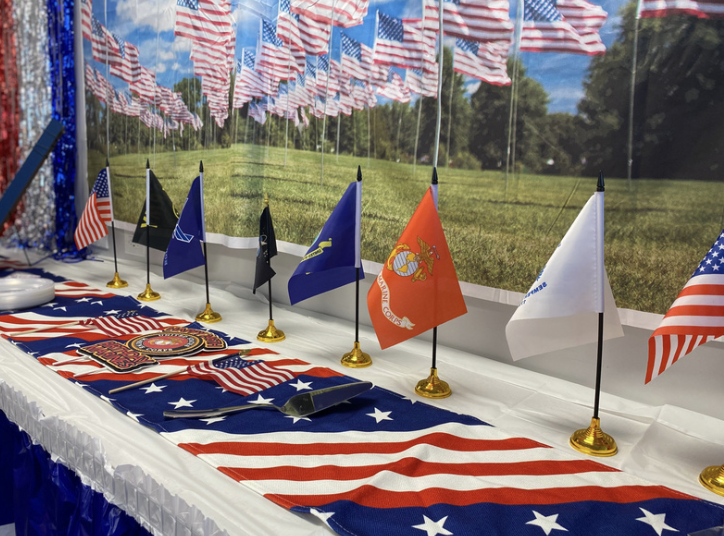 Table decorated with American flag tablecloth, with small American and military flags displayed in a row. The backdrop features a field with multiple American flags. The scene is bright with a sunny sky.