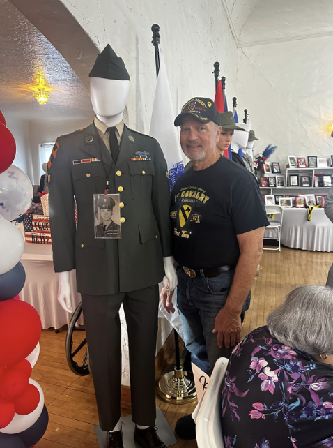 A man wearing a black Veterans shirt and a cap is standing next to a mannequin dressed in a military uniform at a Veterans tribute or memorial event.