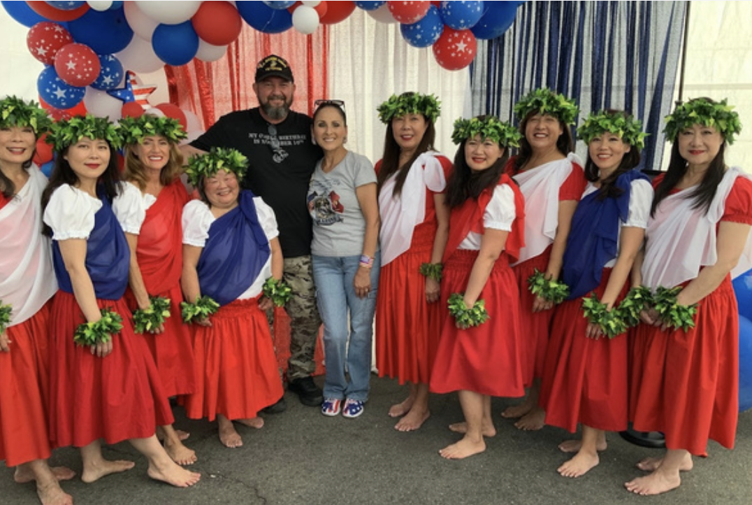 Group of women dressed in traditional Hawaiian hula costumes with red skirts, white tops with red or blue accents, and green leafy headbands and wristlets, standing with two men in casual clothing amidst patriotic decorations of red, white, and blue 