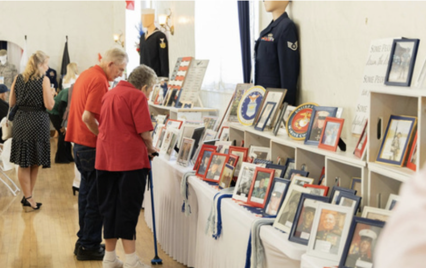 People browsing framed photos and memorabilia at a display table in a museum or gallery setting, with mannequins in military uniforms in the background.
