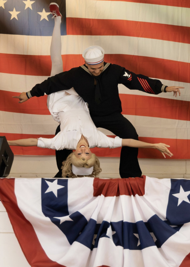 A sailor and a woman performing a choreographed dance or acrobatic pose in front of an American flag backdrop, with patriotic decorations.