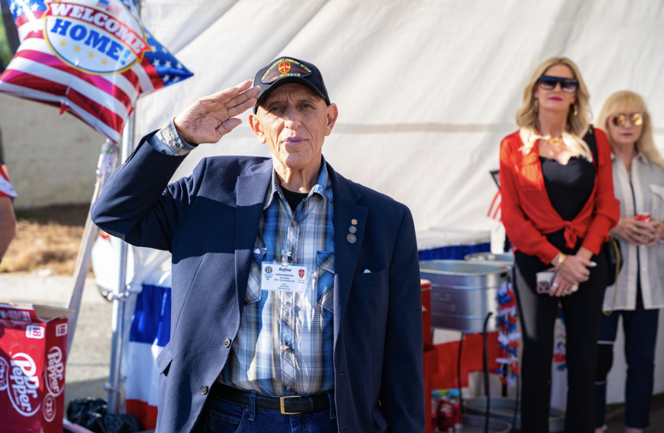 An elderly man saluting at an outdoor event, with a 'Welcome Home' American flag balloon and refreshments in the background. Two women stand behind him, wearing patriotic colors.