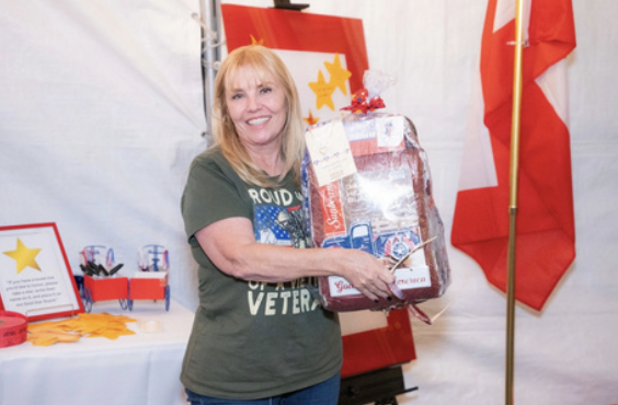 A woman with blonde hair smiling and holding a large gift basket, standing in front of a white tent with red decorations and a Canadian flag.