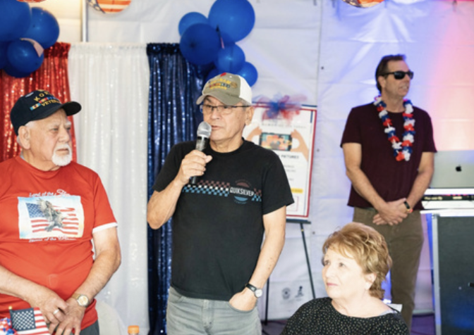 Group of four people at a decorated event, one man wearing a black shirt and cap speaking into a microphone, another man wearing sunglasses and a floral lei, a woman with short hair sitting, and an elderly man with a beard wearing a red shirt and cap
