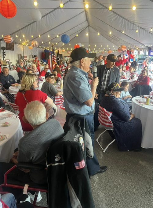 A gathering of older adults at a celebration under a decorated tent, with patriotic and festive decorations including string lights and paper lanterns, and red, white, and blue themed table settings.