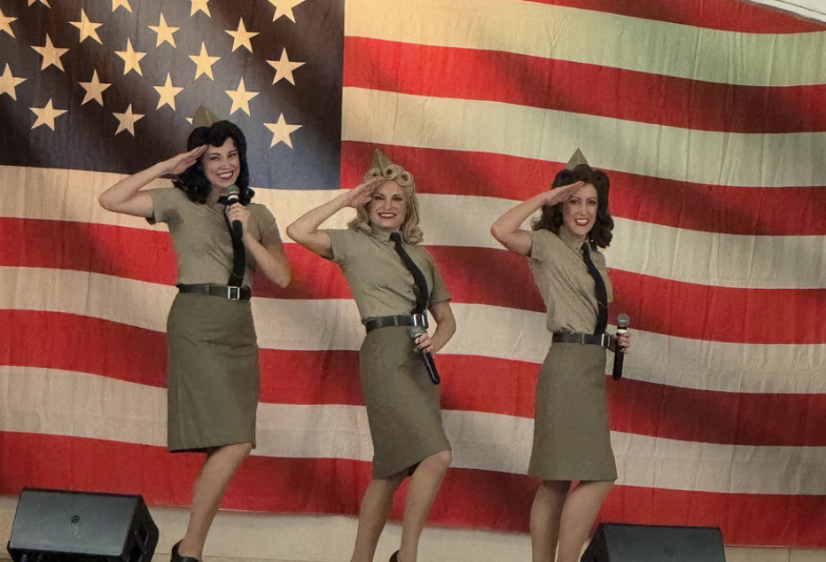 Three women in vintage military-style dresses saluting on stage with an American flag backdrop.