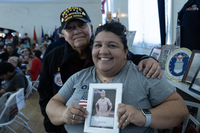 A woman smiling and holding a framed photo of a soldier, with an older man standing close behind her at a gathering honoring veterans.