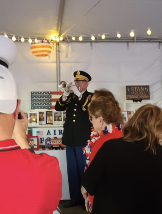 A man in a military uniform playing a trumpet at a patriotic event with an American flag-themed lantern hanging from the ceiling and people watching him.