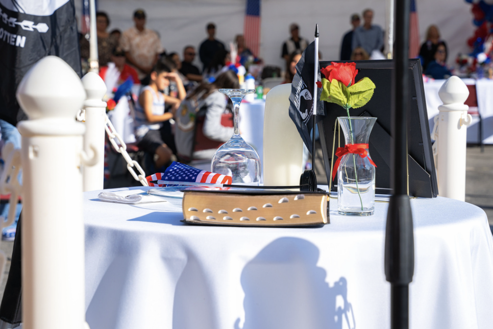A table decorated with patriotic items including a red rose in a glass vase, an American flag, a water glass, and a book, with a patter of patriotic-themed decorations and a crowd of people in the background at an outdoor event.