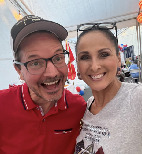 A man and woman smiling and taking a selfie inside a tent at an event. The man is wearing glasses, a baseball cap, and a red shirt. The woman is wearing glasses on her head and a light gray shirt.