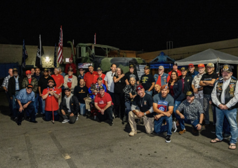 Group of people gathered outdoors at night with military vehicles and flags in the background.